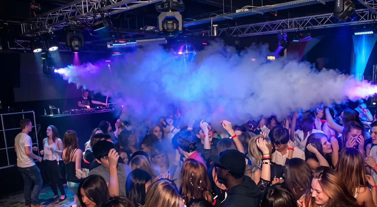 Pista de baile llena de gente frente al DJ, envuelta en niebla iluminada con luces azules y moradas