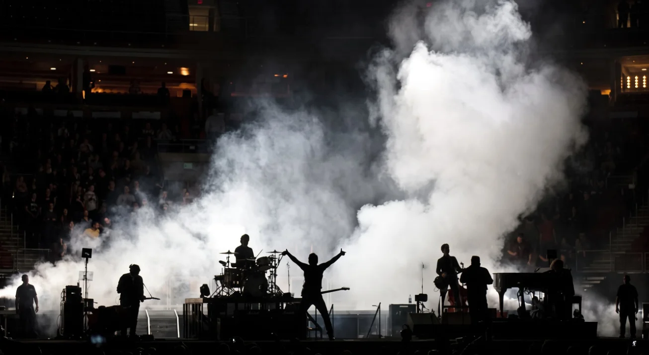 Banda en vivo en un estadio, siluetas de los músicos entre columnas densas de niebla escénica